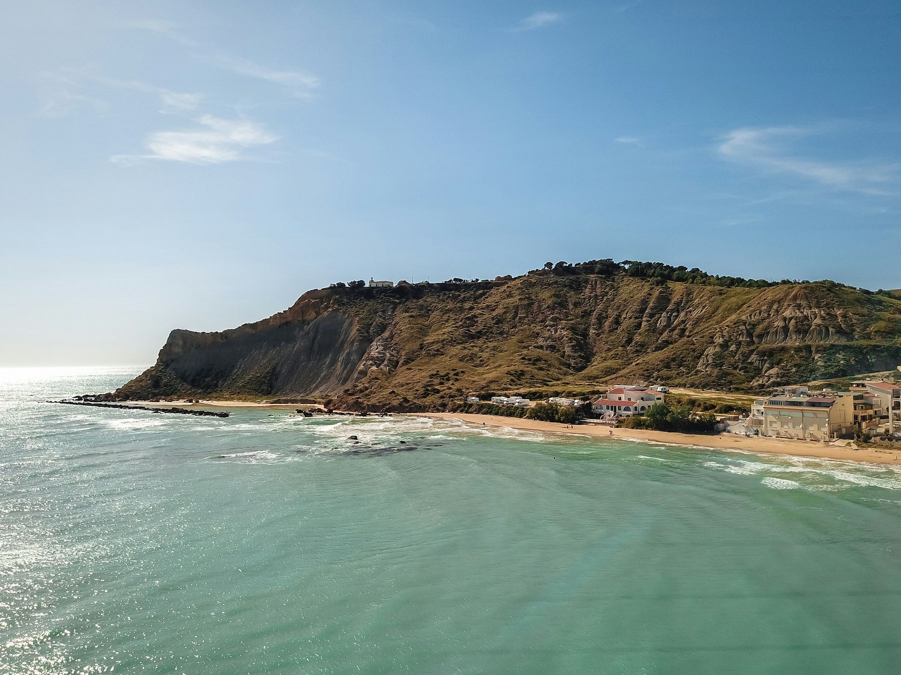 Un promontorio bruno segnato dai calanchi, con un piccolo edificio bianco in cima, sopra una spiaggia con case e palazzine, visto dal mare con un drone