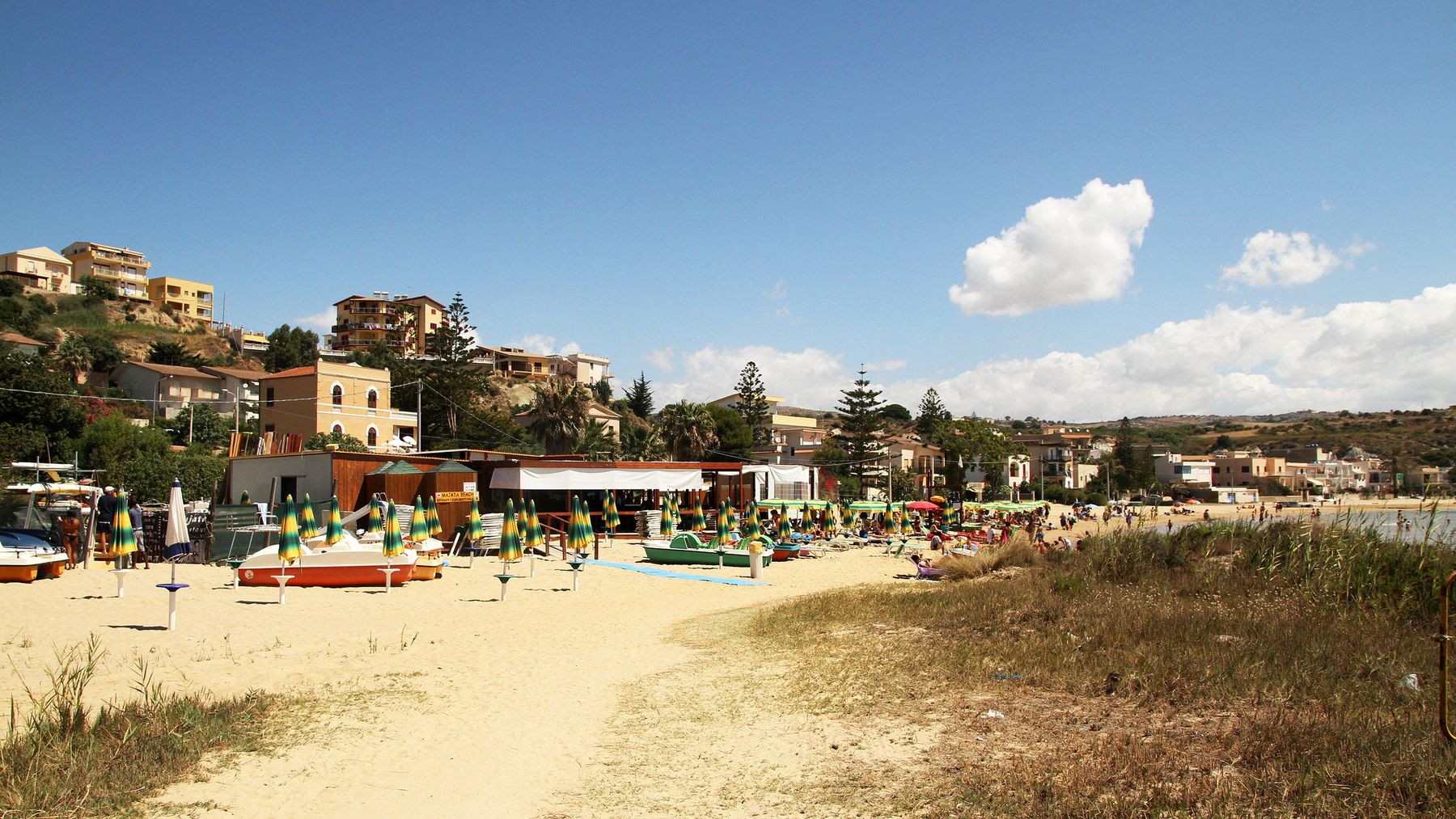 La spiaggia di Punta Grande d'estate: ombrelloni chiusi a strisce verdi e gialle e pedalò sulla sabbia, il chiosco di uno stabilimento con il cartello Majata beach e, dietro, case e palazzine sulla collina