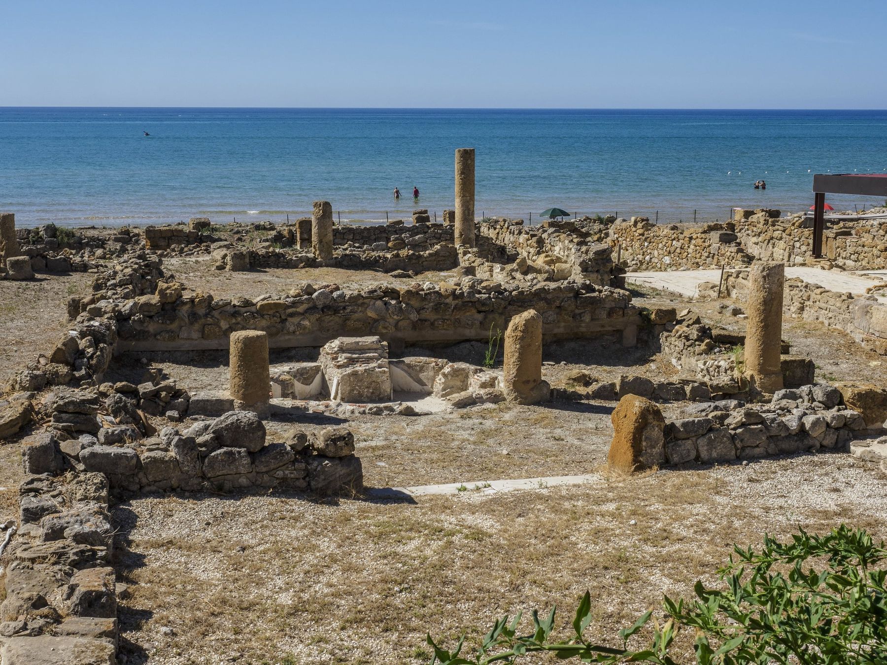 Gli scavi della villa romana di Realmonte: muri bassi di pietra, alcune colonne ancora in piedi e una vasca al centro, con il mare blu e alcuni bagnanti lontani sullo sfondo