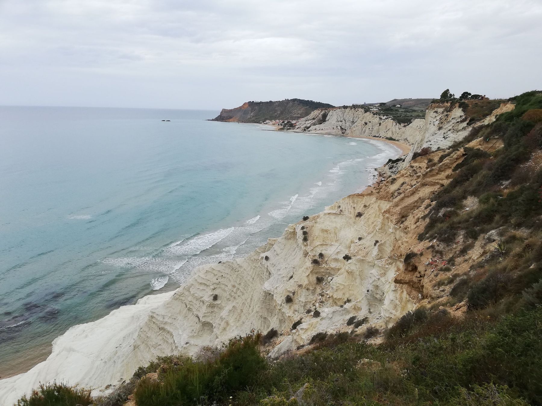 Dal belvedere in alto: la costa che curva verso ovest con il mare turchese, la falesia di marna chiara in basso e un cielo coperto