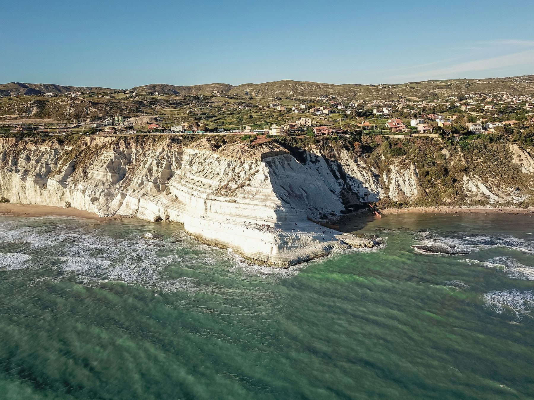 La Scala dei Turchi vista dal mare con un drone: la parete di marna bianca affiora sull'acqua verde con le onde, e alcune case sul ciglio della falesia