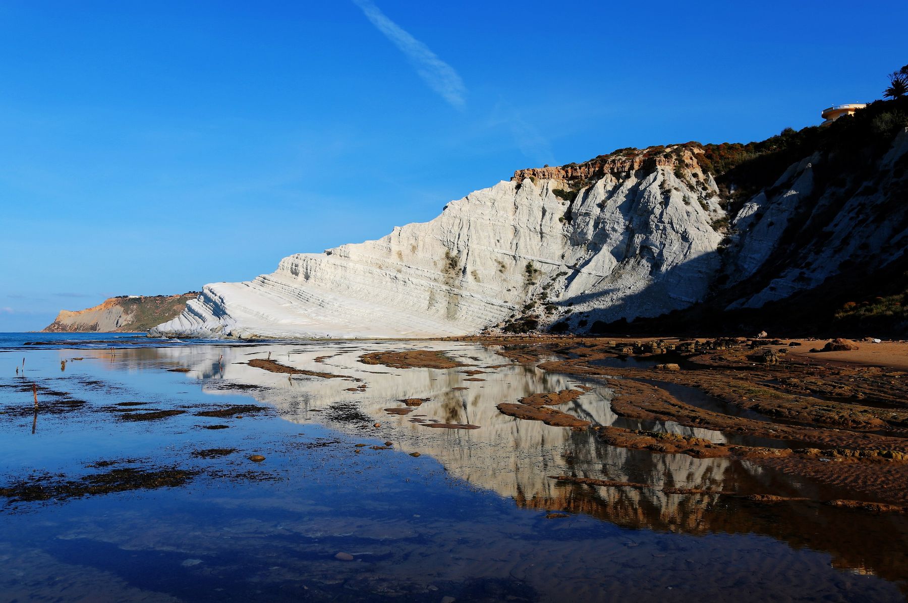 La scogliera di marna bianca della Scala dei Turchi con i gradoni, vista dalla battigia nella luce del mattino, riflessa nelle pozze di mare sotto un cielo blu intenso