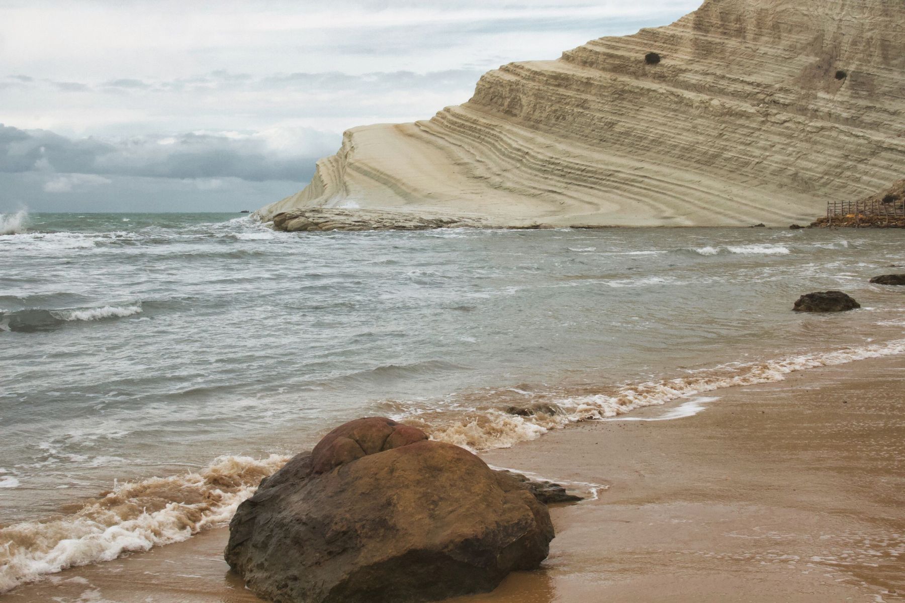 La Scala dei Turchi in una giornata di vento: mare mosso e cielo coperto, un grande masso sulla sabbia bagnata in primo piano e la marna color crema sullo sfondo