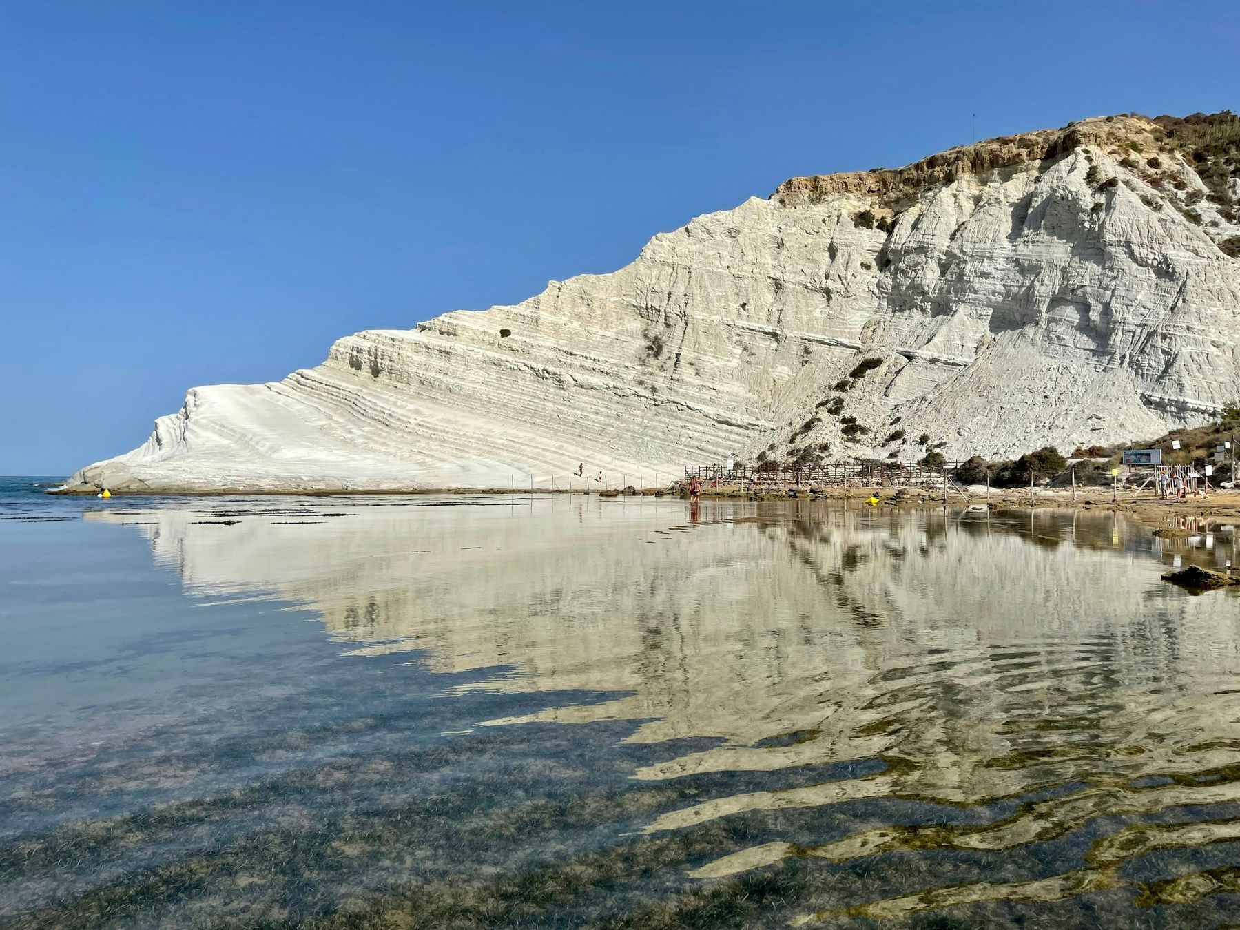 La Scala dei Turchi dal bagnasciuga, con il mare liscio che ne riflette i gradoni bianchi e, a destra, la passerella di legno del lido