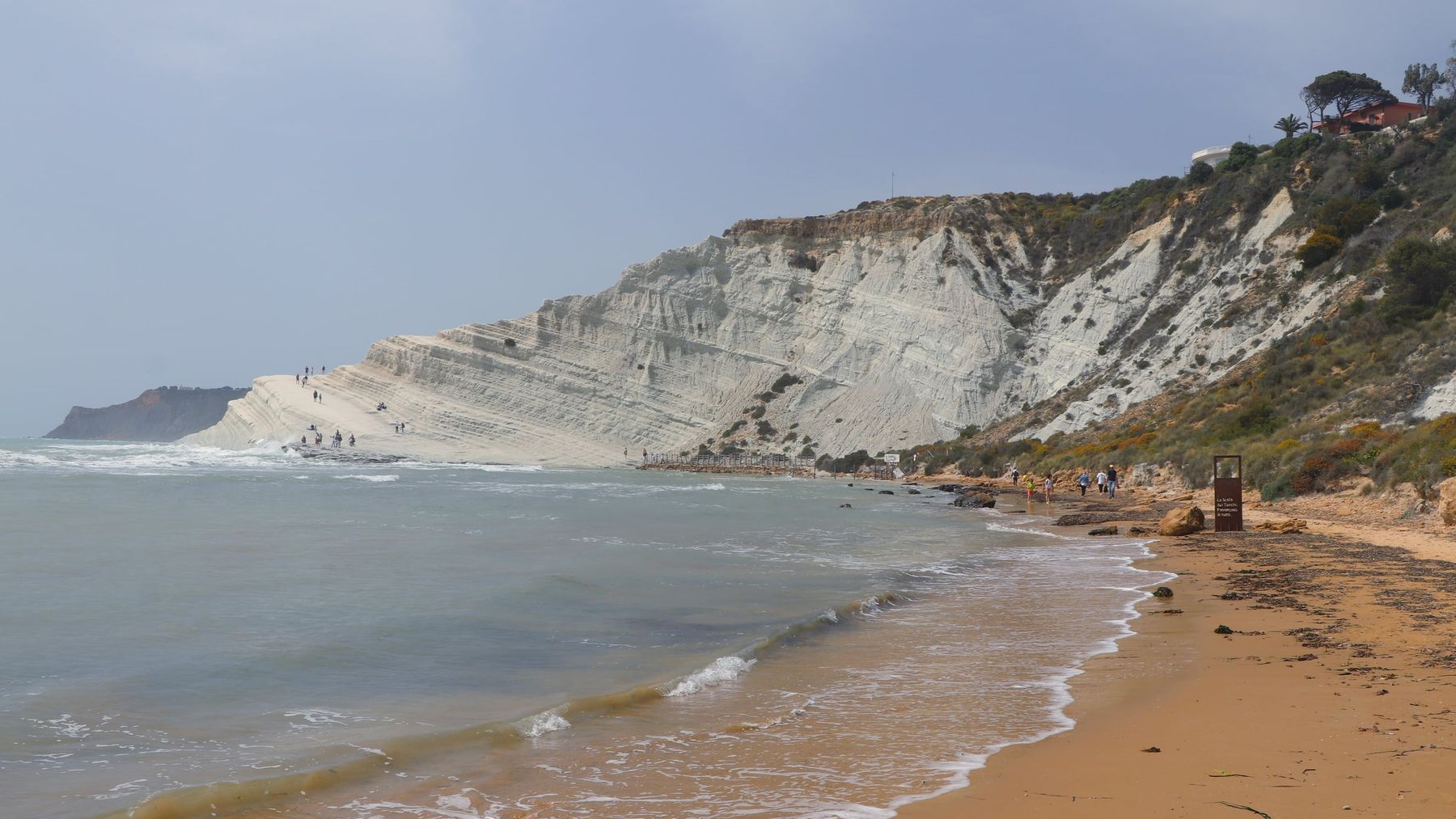 Dalla spiaggia di sabbia rossiccia si vede in lontananza la Scala dei Turchi, con persone minuscole sui gradoni e alcuni escursionisti che camminano lungo la riva sotto un cielo carico