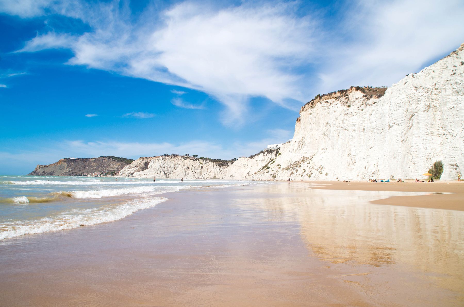 La spiaggia bagnata ai piedi della falesia di marna bianca, con le onde che arrivano a riva, nuvole alte nel cielo azzurro e pochi bagnanti in lontananza