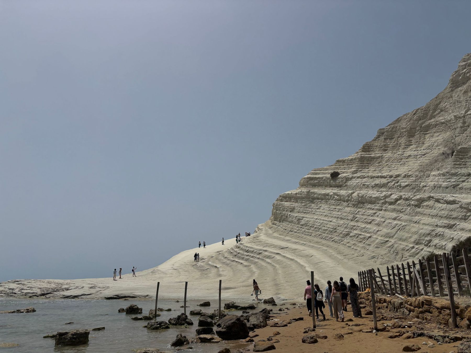 Un gruppo di visitatori cammina sulla sabbia accanto alla recinzione di pali e rete di legno ai piedi della Scala dei Turchi, mentre altre persone sono sui gradoni più in alto