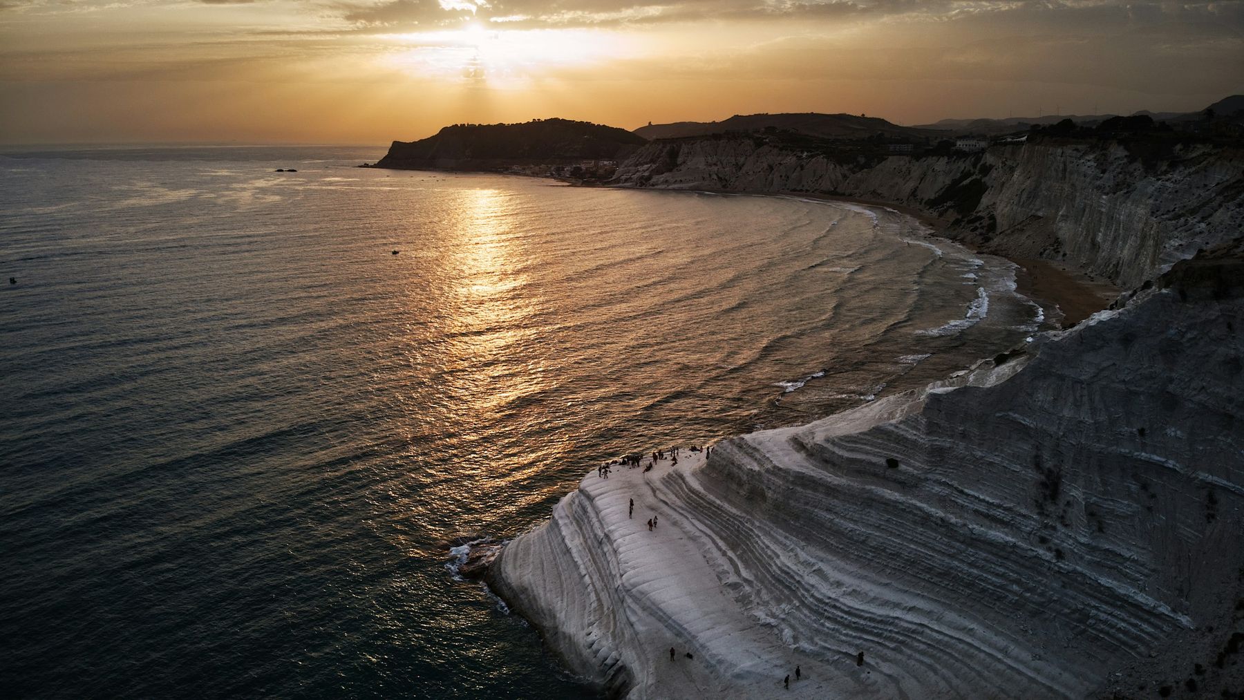 Veduta aerea al tramonto: il sole cala dietro un promontorio e lascia una scia dorata sul mare, mentre sui gradoni bianchi in primo piano si vedono piccole figure