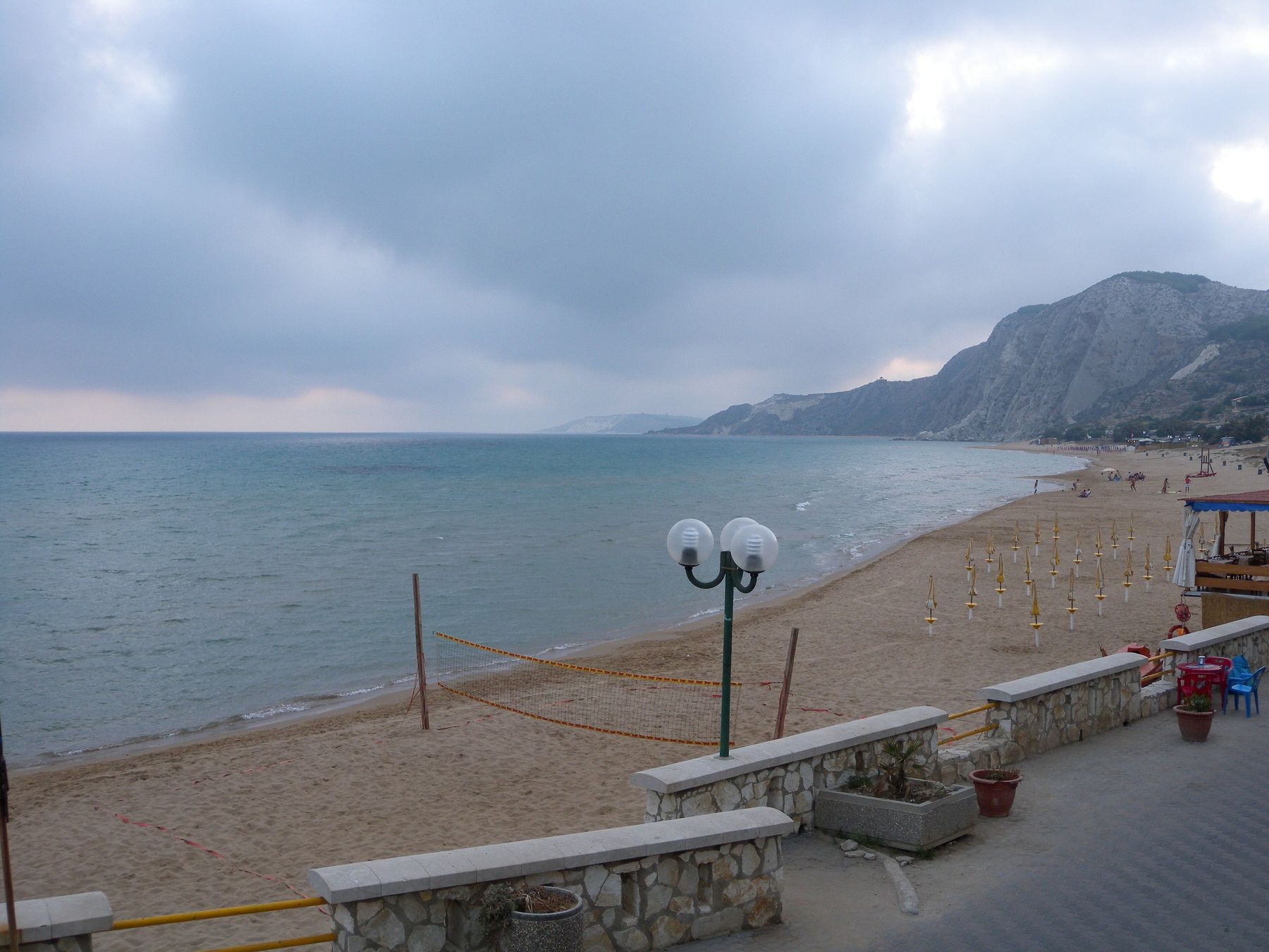 Die Strandpromenade von Siculiana Marina an einem bewölkten Abend: vorn eine niedrige Steinmauer und eine Laterne, der Sandstrand mit einem Volleyballnetz und geschlossenen Sonnenschirmen und ein heller Hügel, der zum Meer abfällt